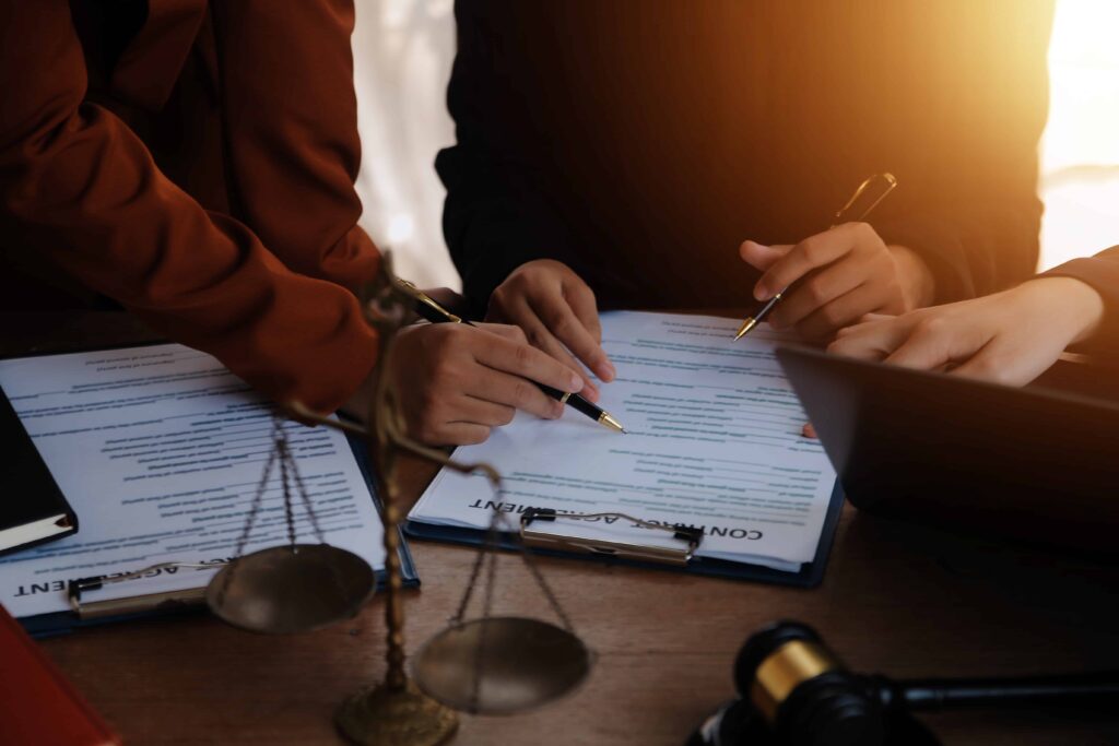Lawyers reviewing and signing legal contract documents at a desk with scales of justice and gavel.